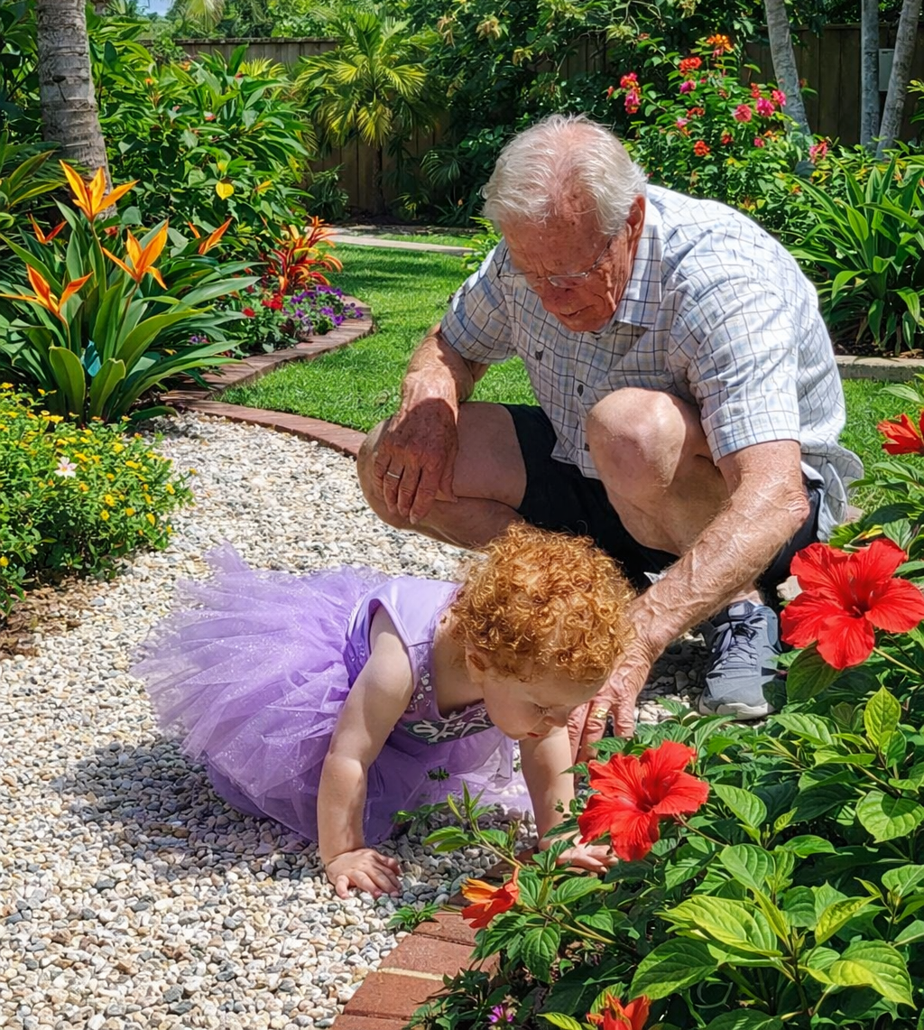 A grandfather crouching in a lush garden with a curly-haired toddler in a purple tutu — multigenerational wonder at Porchaven™