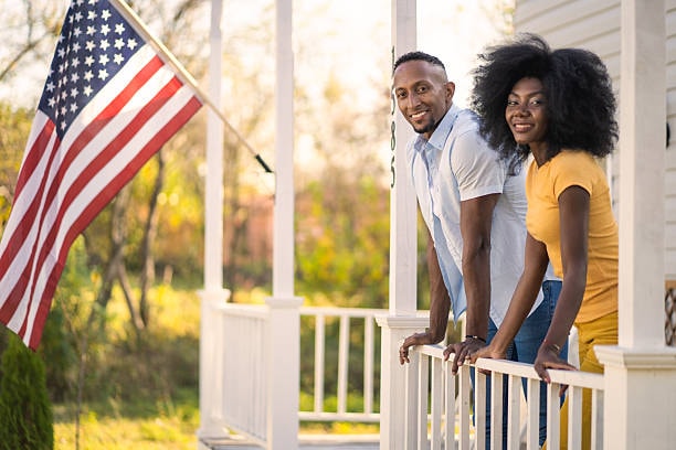 A couple smiling on their front porch with an American flag — the warmth of front-porch community living